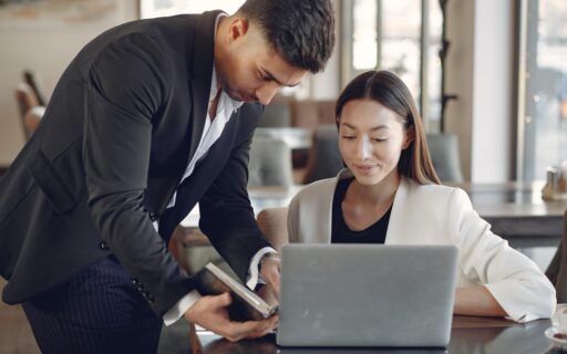 Male hovering over female showing her a book while she is on a laptop
