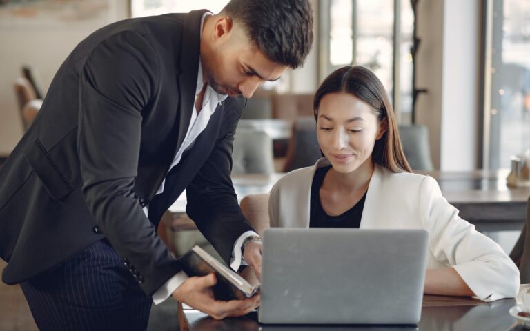 Male hovering over female showing her a book while she is on a laptop